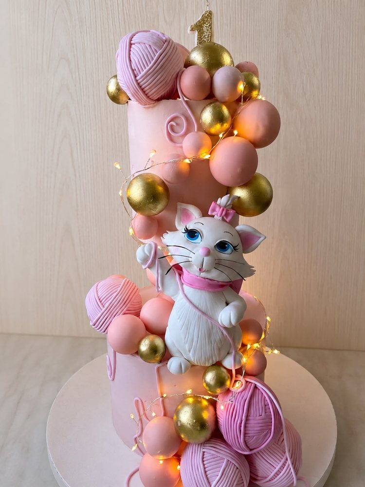 Roze rođendanska torta sa figurom mace i zlatnim kuglama.